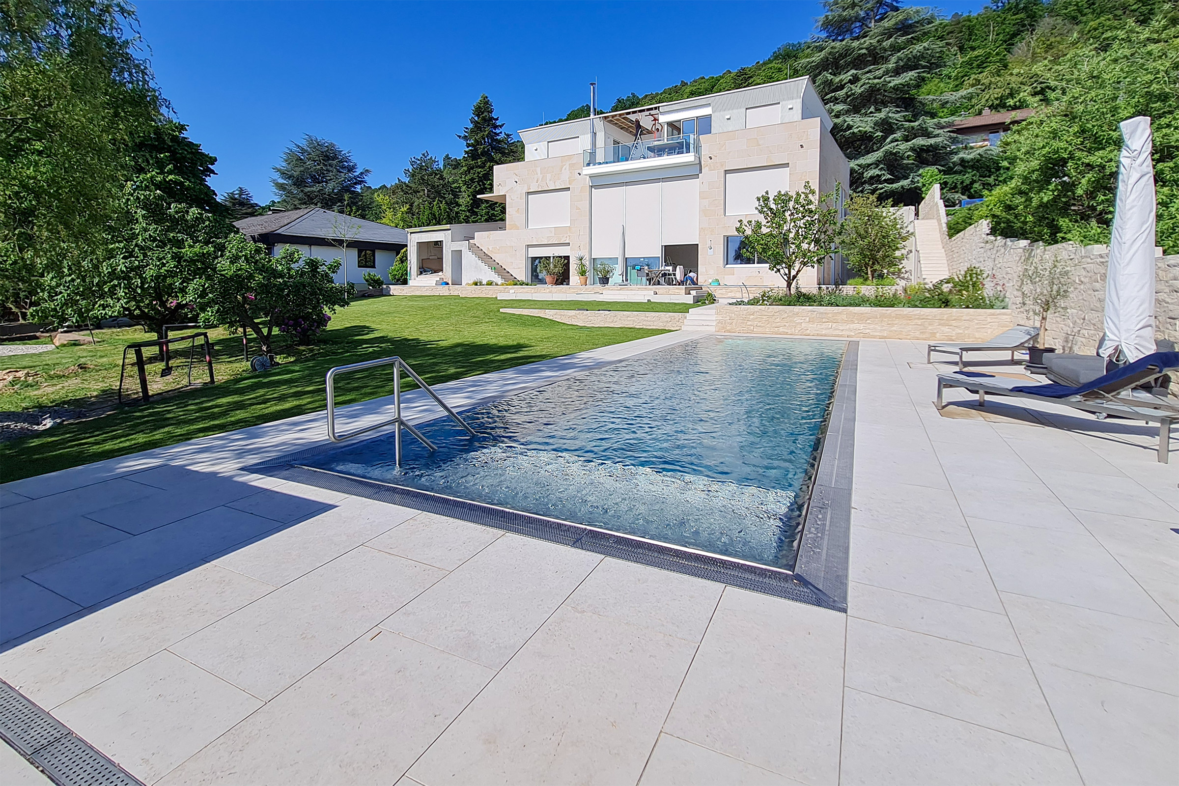 Edelstahlpool mit Luftsprudelliege und moderner Architektur im Hintergrund Edelstahlpool mit Luftsprudelliege, gerader Treppe und Blick auf eine moderne Villa mit Gartenanlage