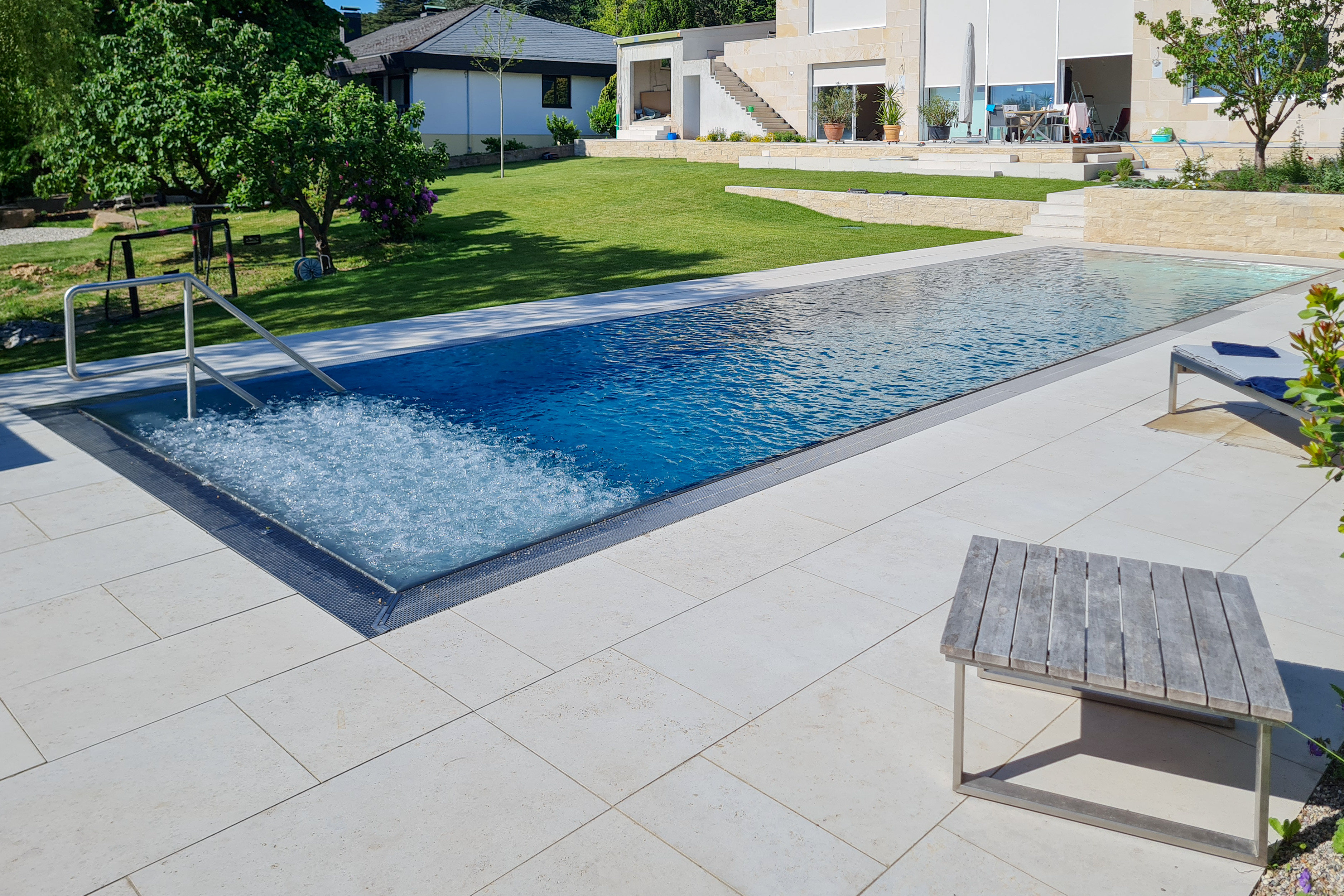 Edelstahlpool mit Überlaufrinne und Luftsprudelliege in Betrieb Langgezogener Edelstahlpool mit Luftsprudelliege auf voller Breite und Blick auf den Garten mit moderner Außengestaltung