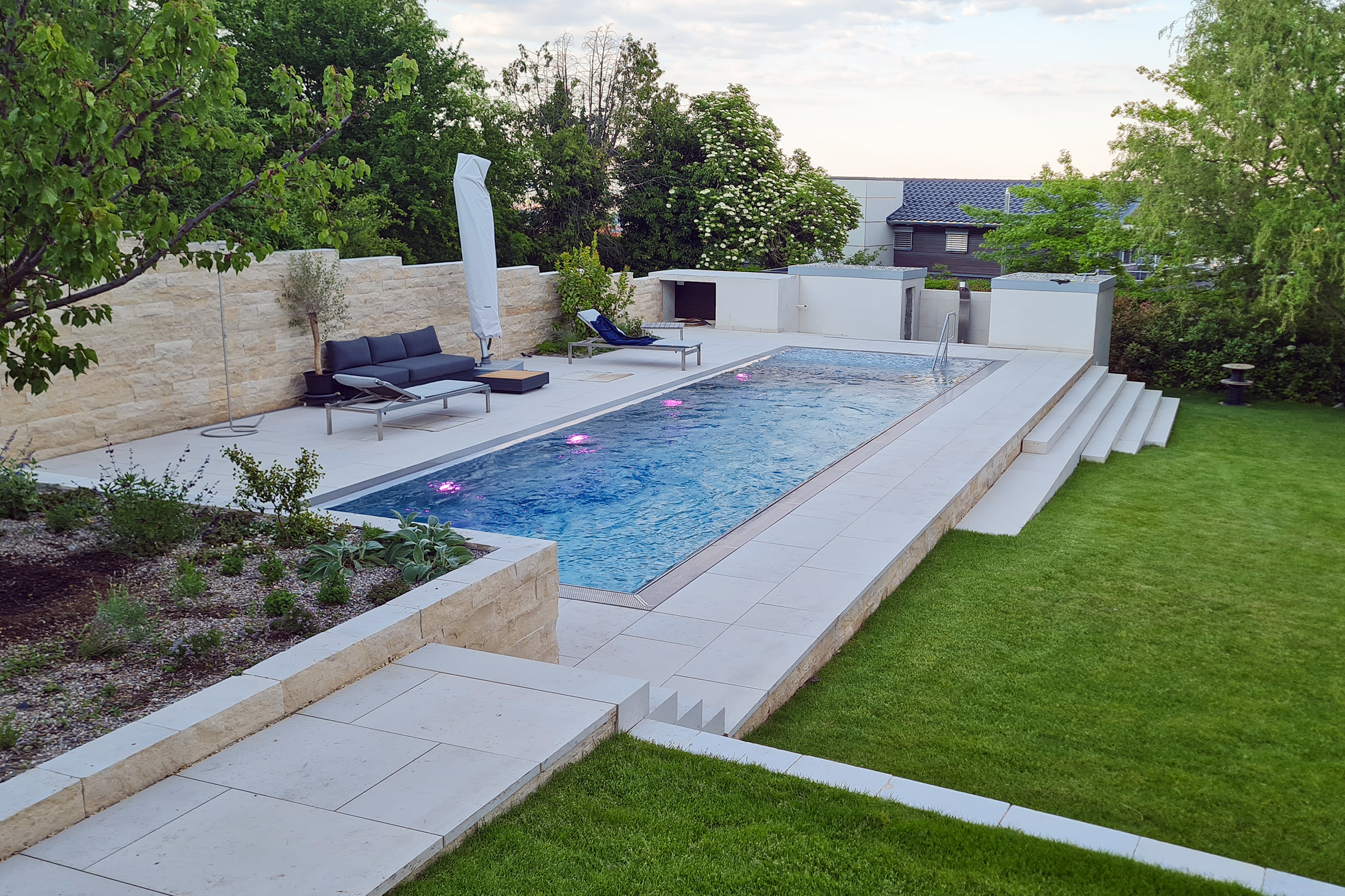 Edelstahl-Überlaufbecken mit eingeschalteter LED-Beleuchtung in der Dämmerung, umgeben von Sitzlounge und Gartenmauer Edelstahlpool am Abend mit beleuchteter Wasserfläche