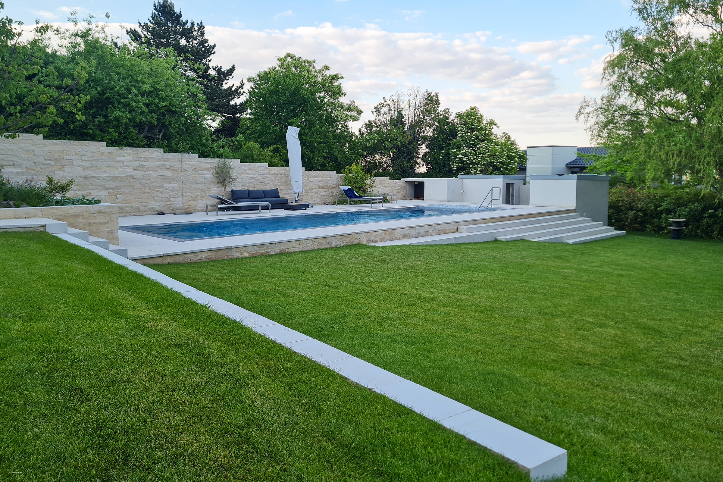Edelstahlschwimmbecken in Gartenterrasse eingebettet Edelstahlpool als Überlaufbecken mit breitem Einstiegsbereich und großzügigem Gartenumfeld mit Natursteinmauer