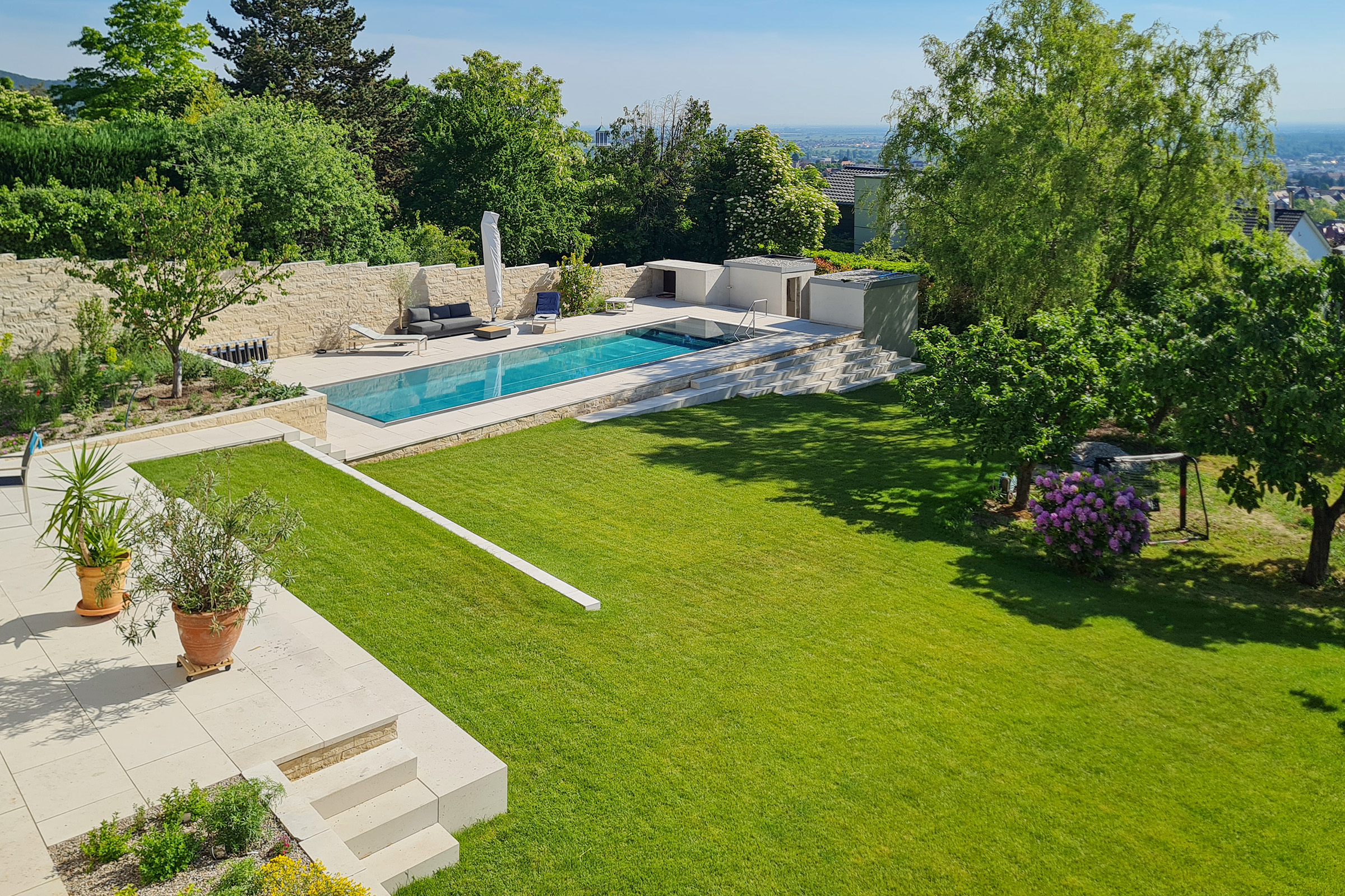 Edelstahl-Überlaufpool mit Stadtblick und großzügiger Gartenfläche Länglicher Edelstahlpool mit gerader Treppe und Blick über die Stadt, umgeben von Grünflächen und Terrasse