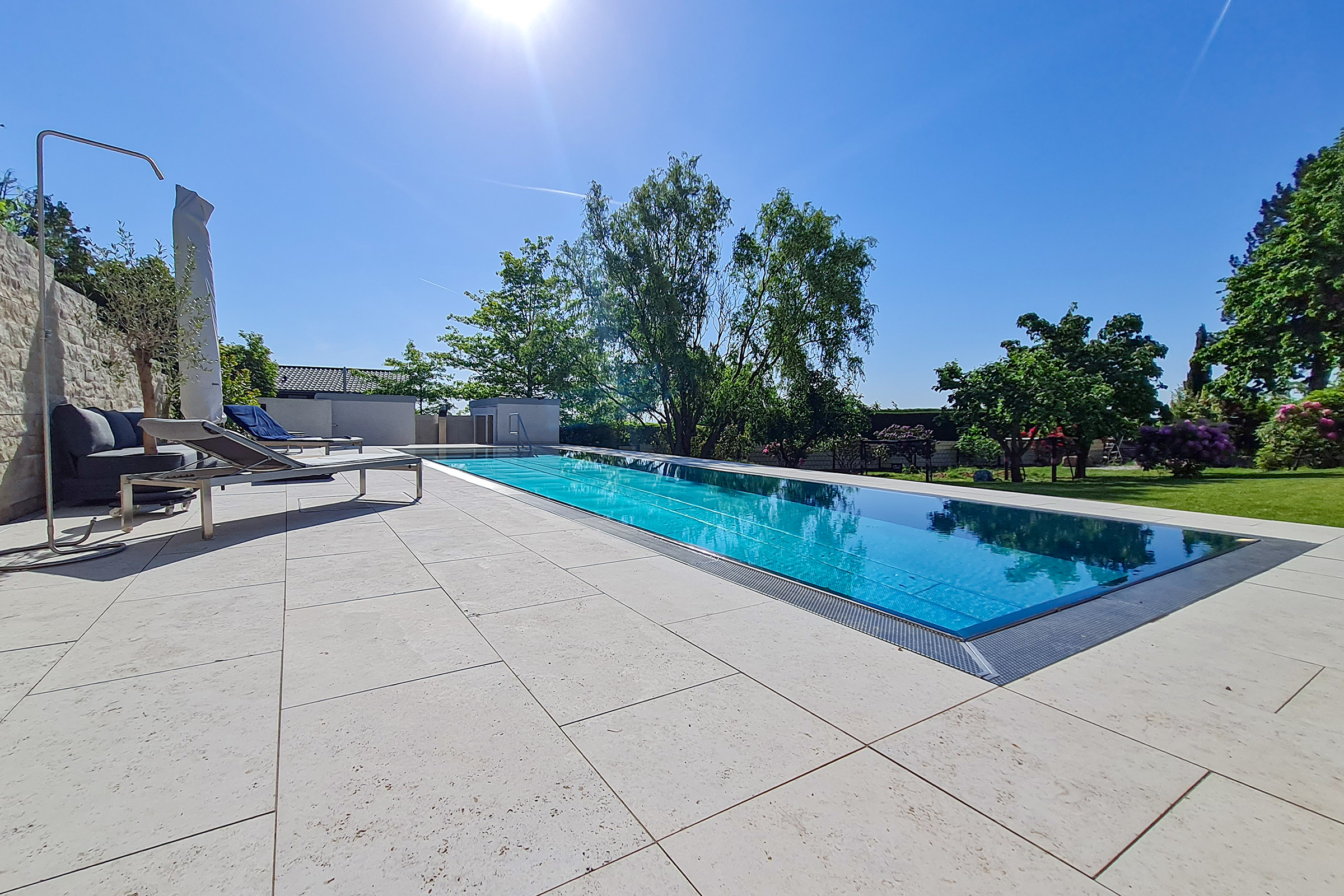 Edelstahlpool mit Rollladenabdeckung bei tief stehender Sonne Flacher Edelstahl-Überlaufpool mit reflektierender Rollladenabdeckung in der Nachmittagssonne, Garten und Lounge im Hintergrund