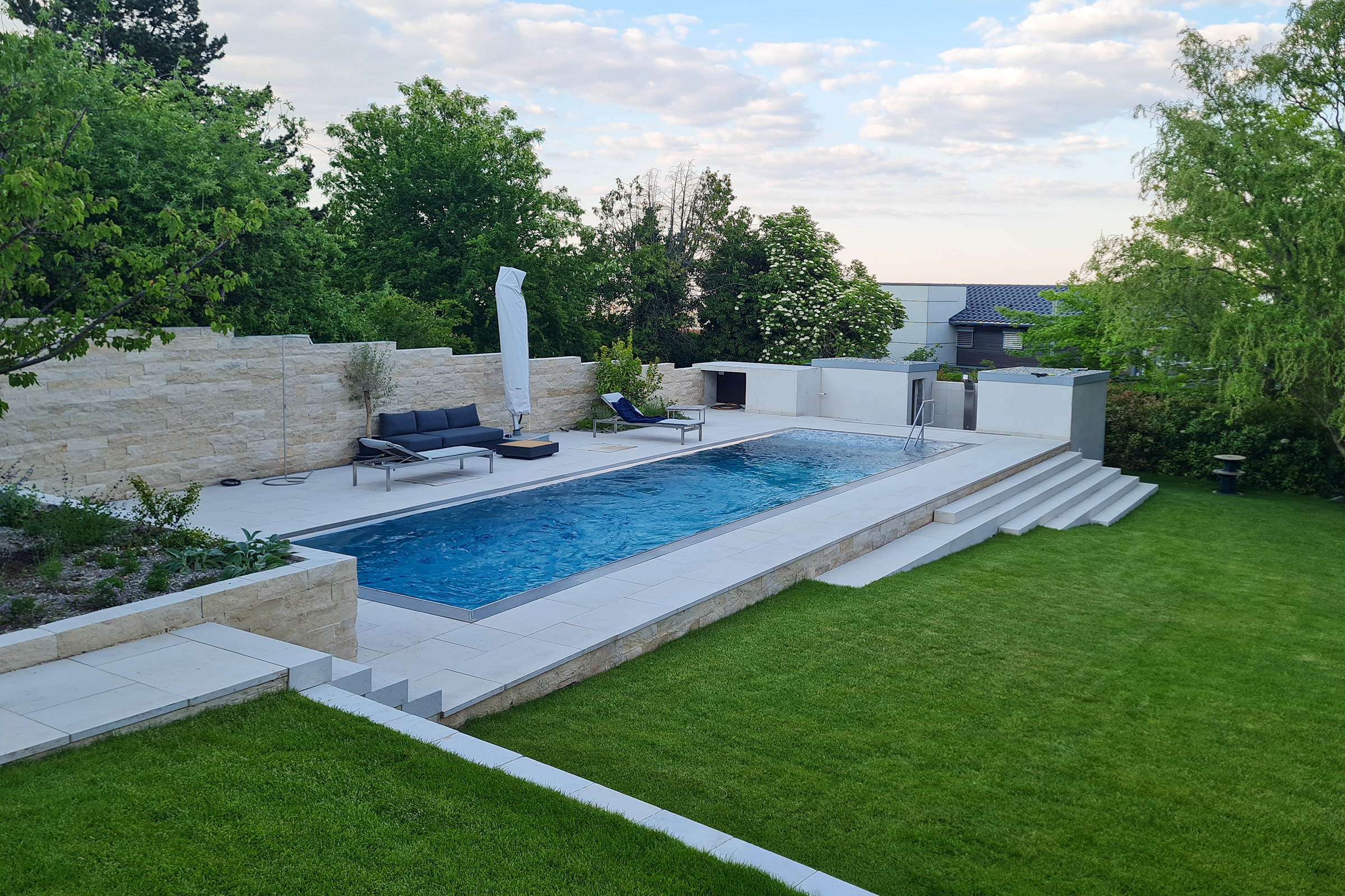 Edelstahlpool eingebettet in moderne Gartenanlage mit Natursteinmauern Großzügiges Edelstahlschwimmbecken mit Überlaufrinne und Sitzecke, eingefasst von Natursteinmauern und Gartenterrassen