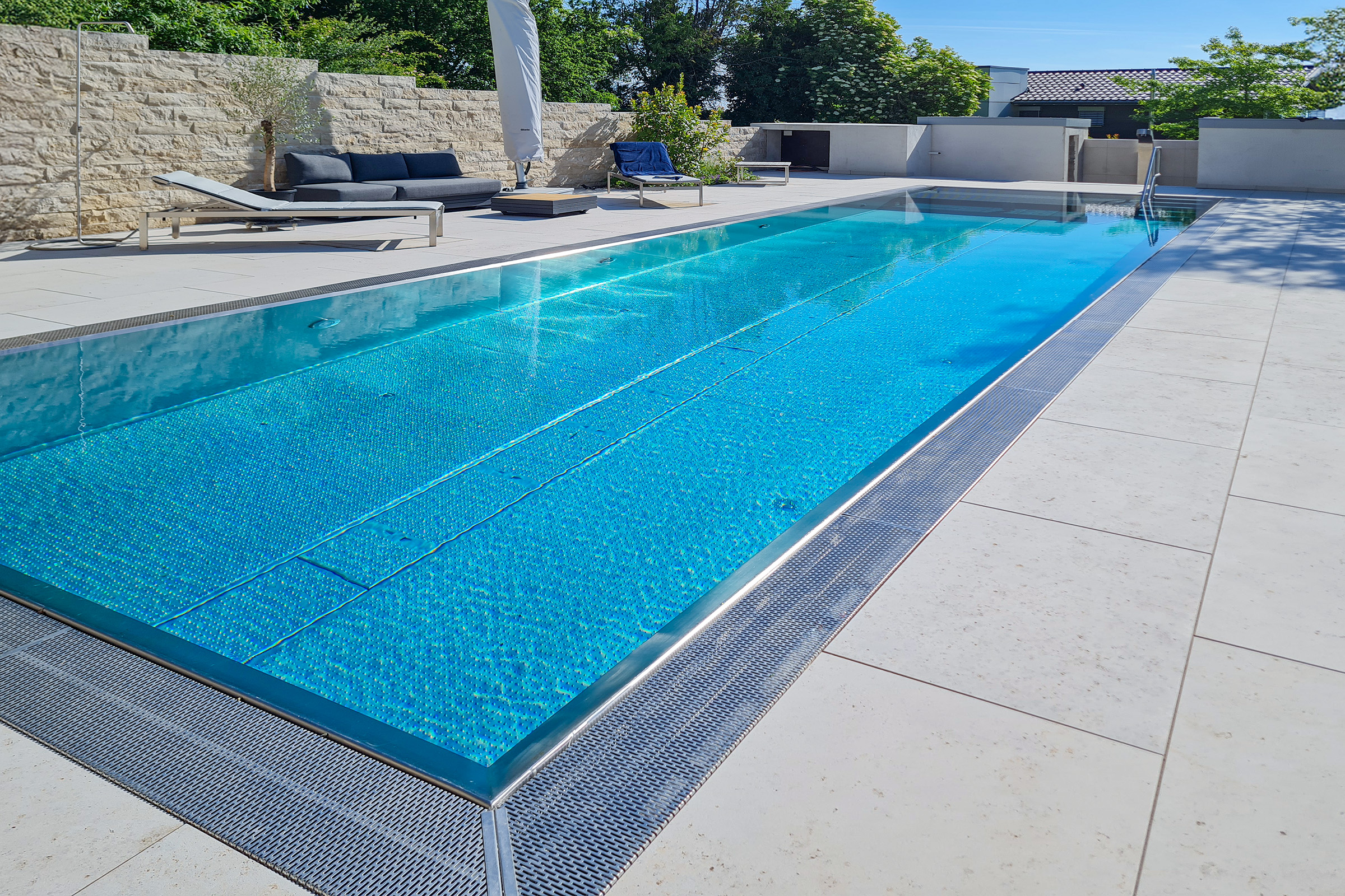 Edelstahlpool mit Rollladenabdeckung im Tageslicht Rechteckiger Edelstahl-Überlaufpool mit Unterflur-Rollladenabdeckung aus Polycarbonatlamellen und angrenzendem Loungebereich
